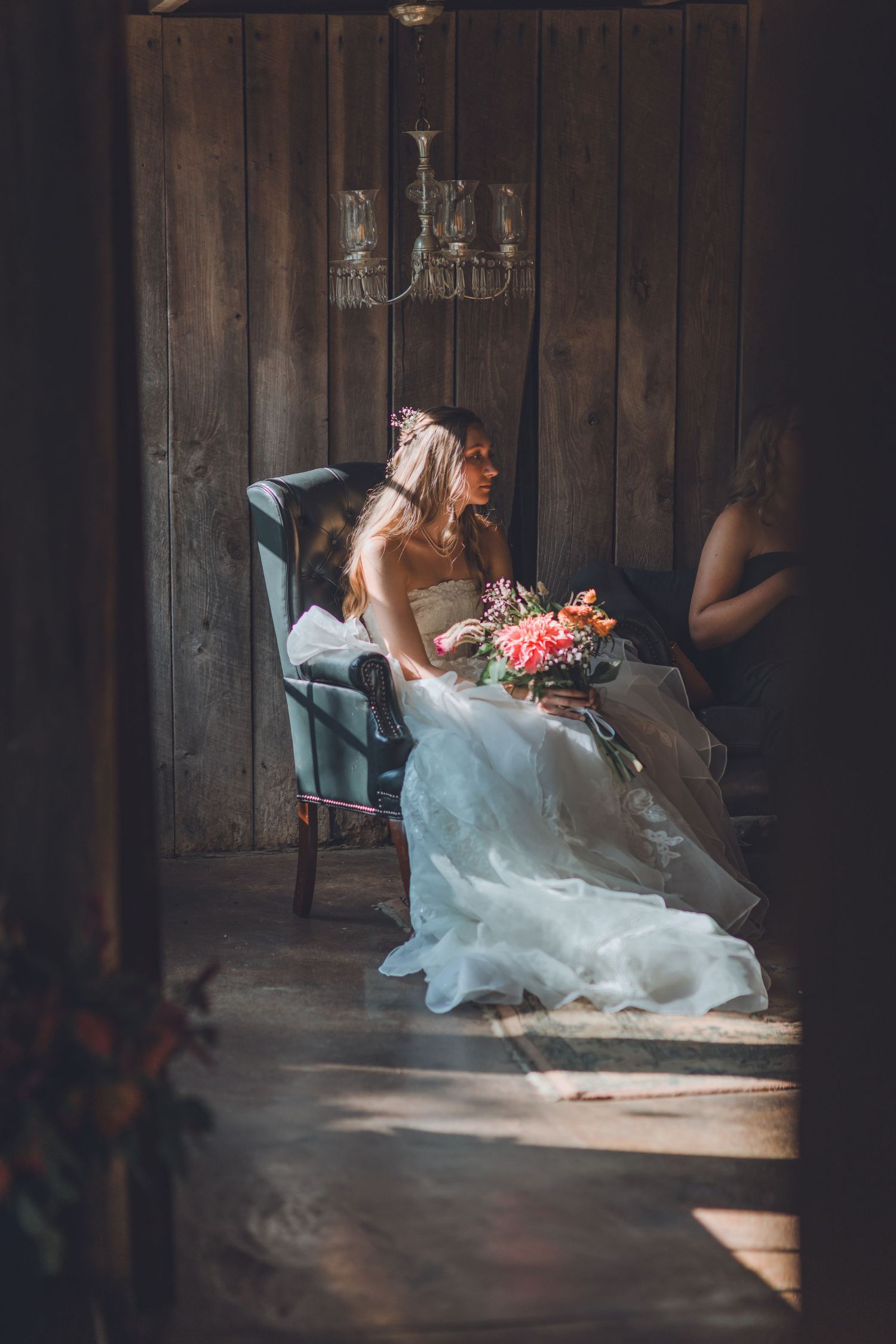 Bride in white gown holding colorful bouquet seated in vintage chair inside rustic wooden barn venue.