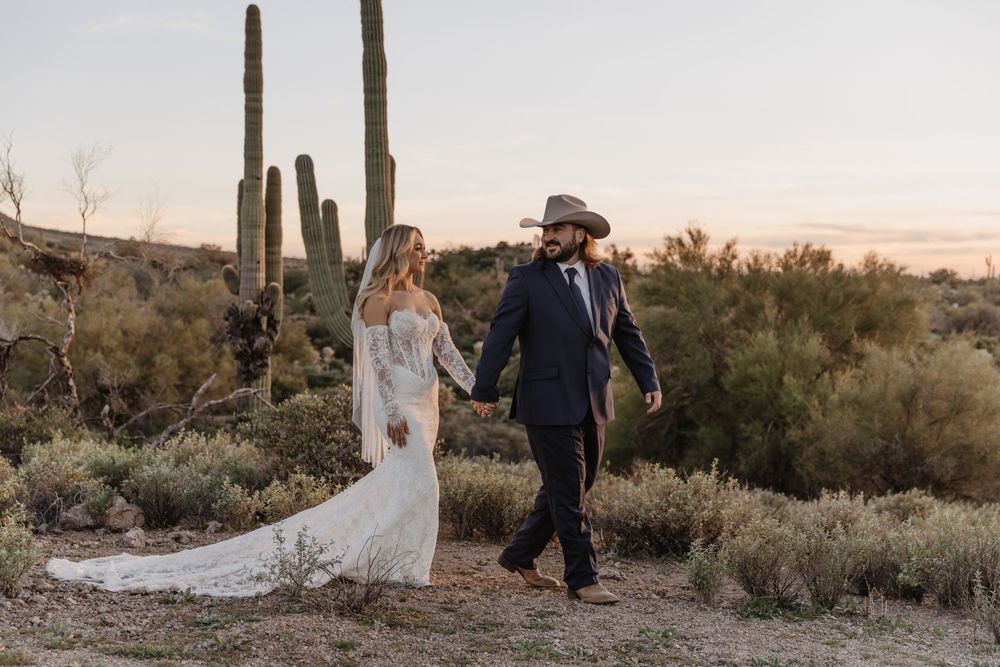 Bride and groom walk hand-in-hand through Arizona desert at sunset, with saguaro cacti backdrop, she in lace gown, he in cowboy hat.