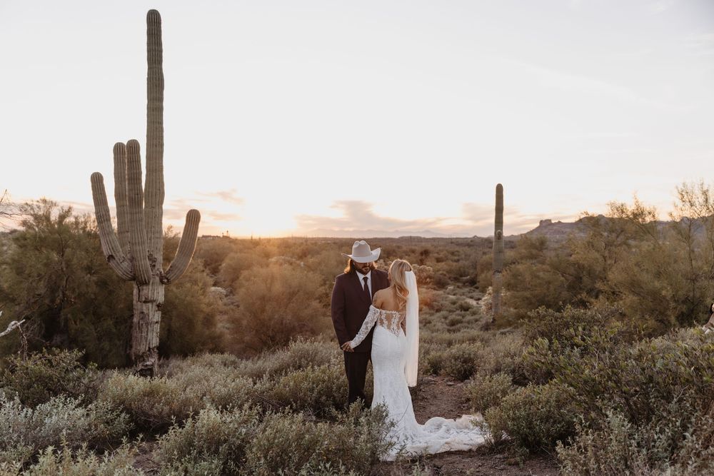 Bride and groom share a romantic moment in the Arizona desert at sunset, with towering saguaro cacti in the background.