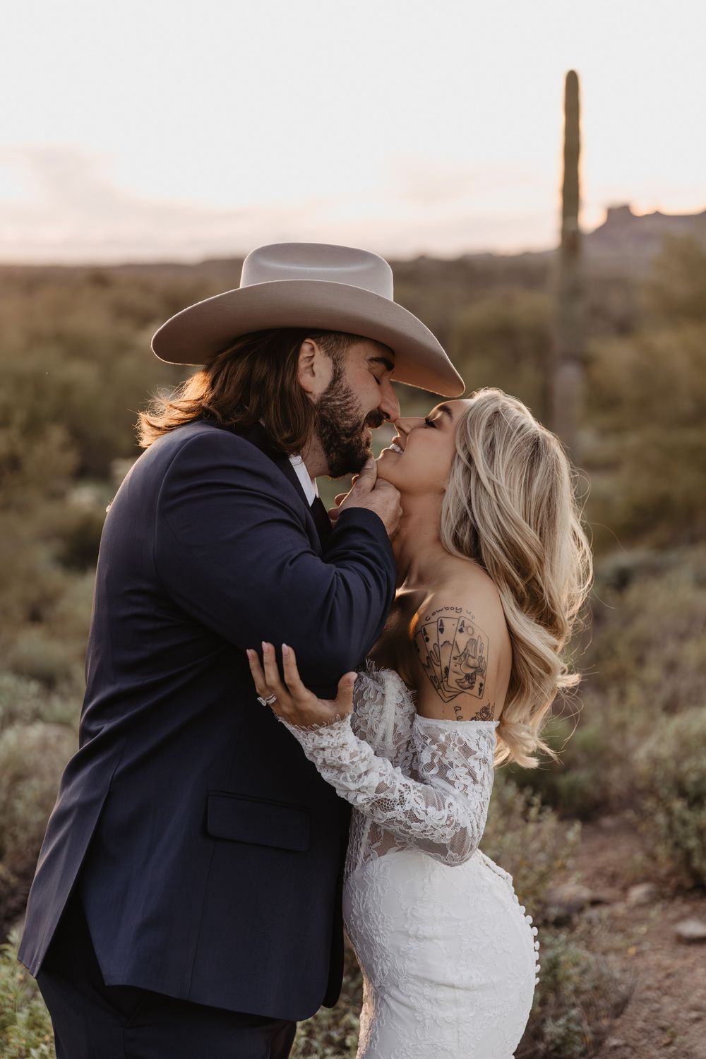 Couple sharing a romantic kiss in the desert at sunset, groom in navy suit and cowboy hat, bride in lace wedding dress.