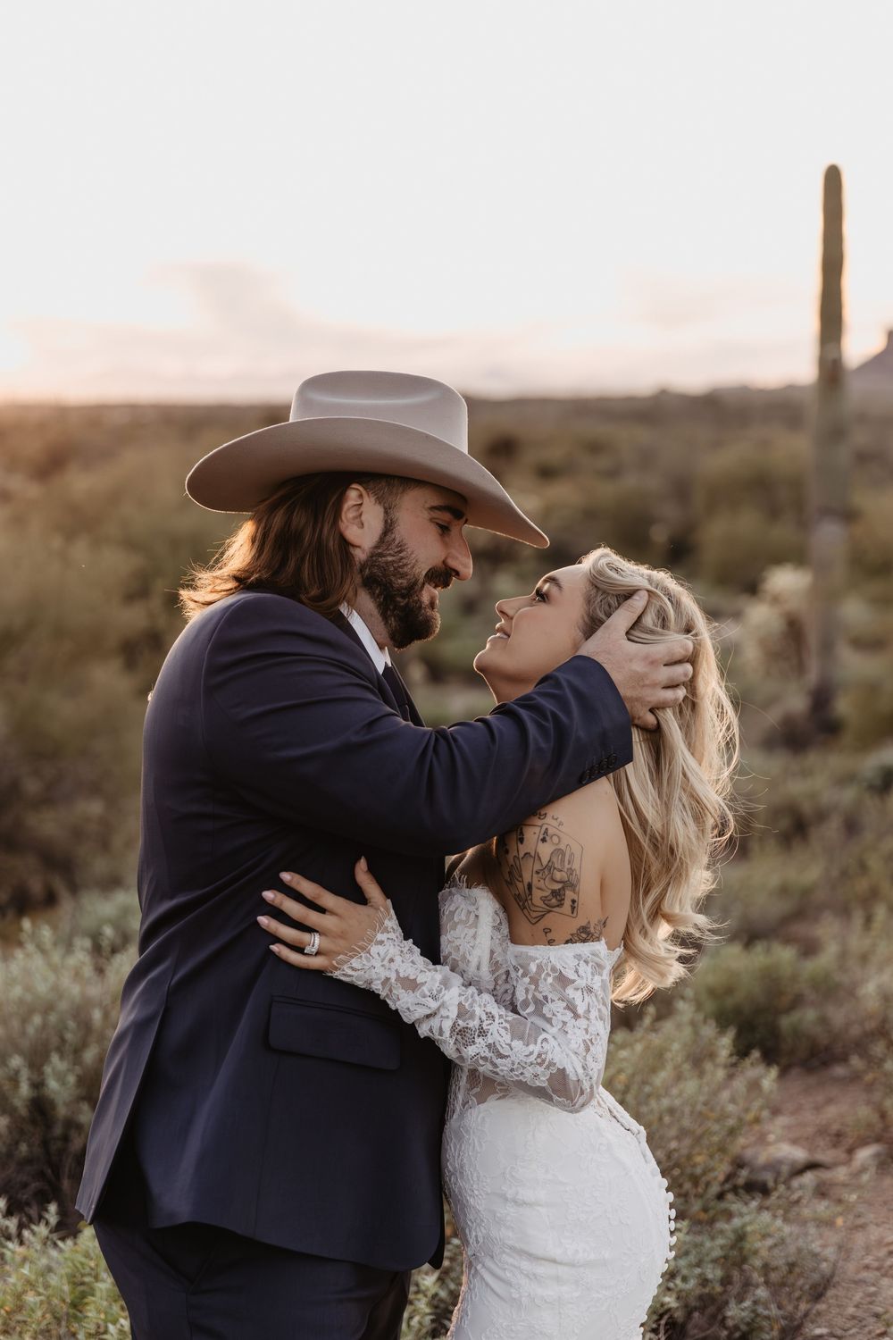 A couple shares a romantic embrace in the Arizona desert at sunset, groom in a cowboy hat and suit, bride in a lace gown.