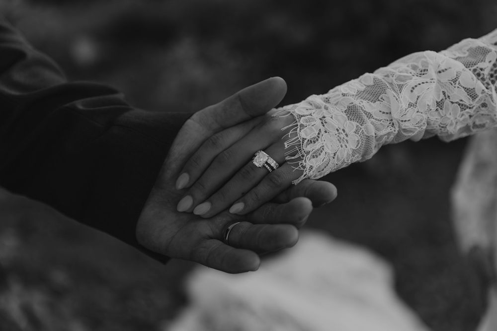 Black and white photo of a bride and groom holding hands, showcasing wedding rings and lace bridal sleeve.