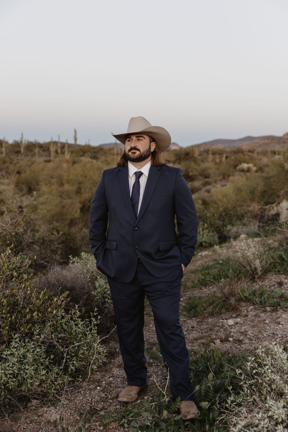 Man in navy suit and cowboy hat stands in desert landscape with saguaro cacti at dusk.