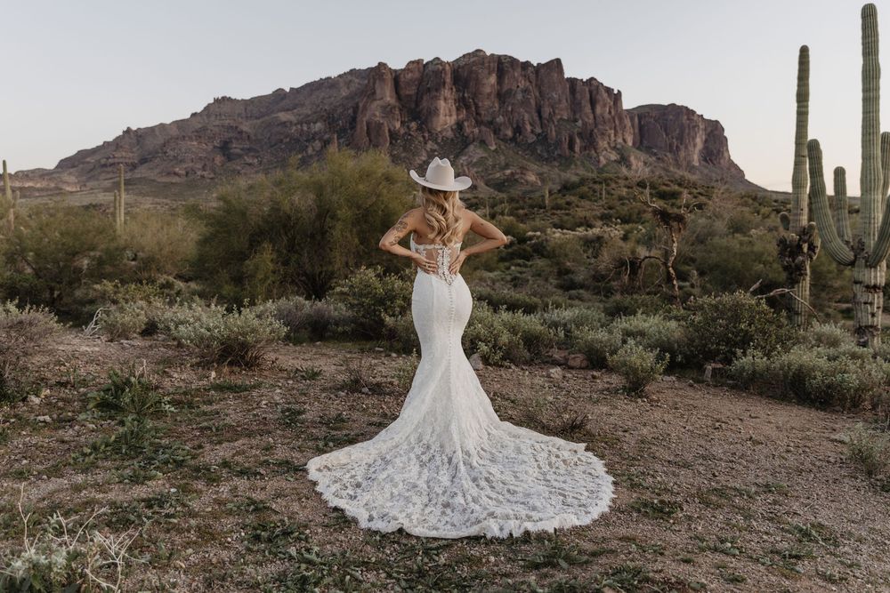 Bride in lace mermaid gown and cowboy hat poses before Superstition Mountains in Arizona desert.