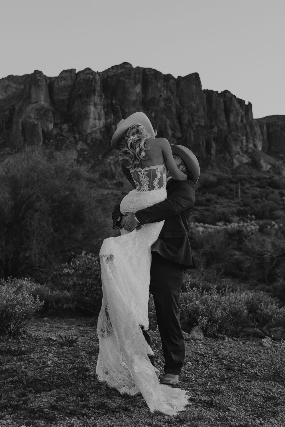 Black and white photo of a couple in cowboy hats embracing before a rocky desert mountain landscape.