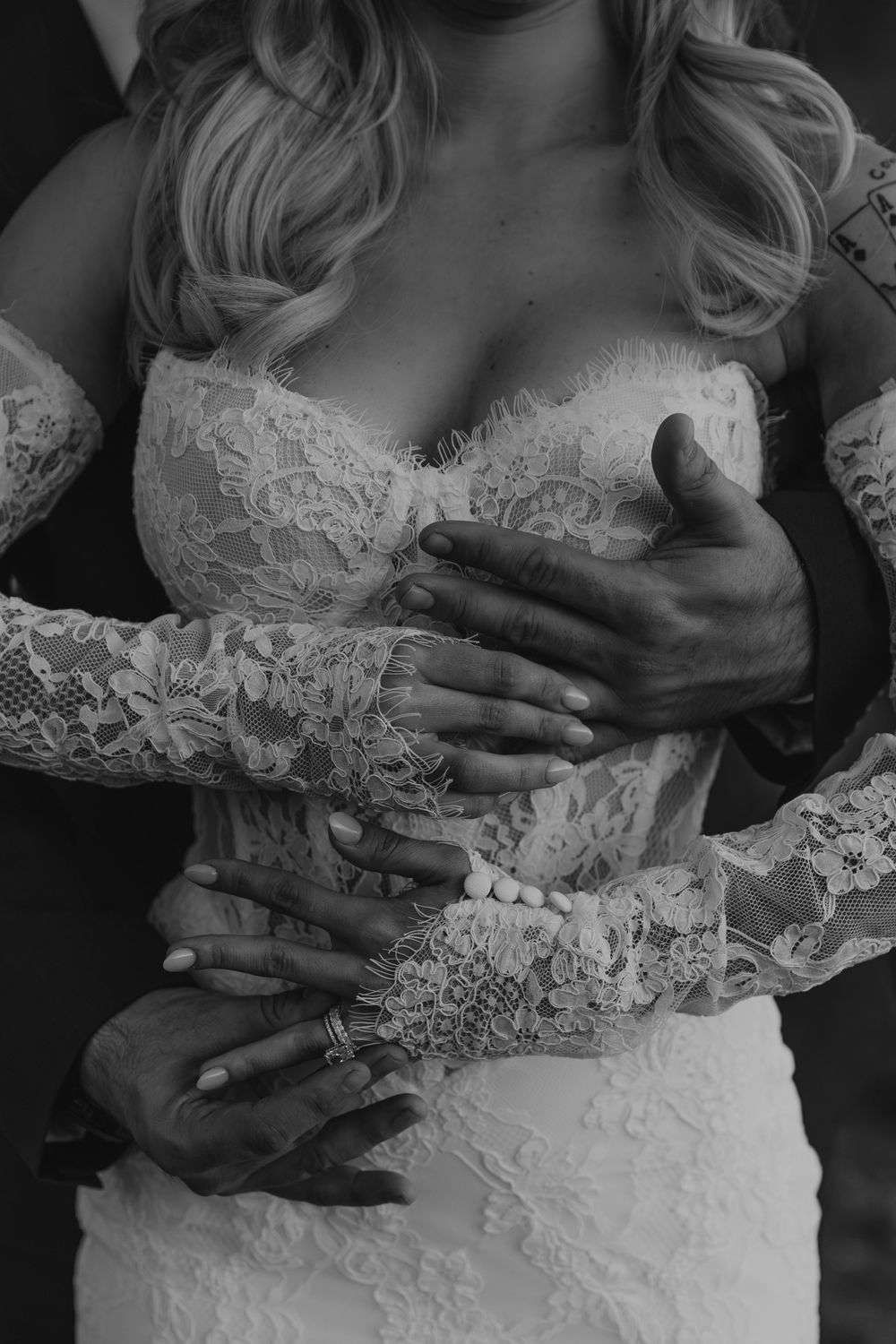 Black and white photo of a bride in lace wedding dress with groom's hands embracing her waist.