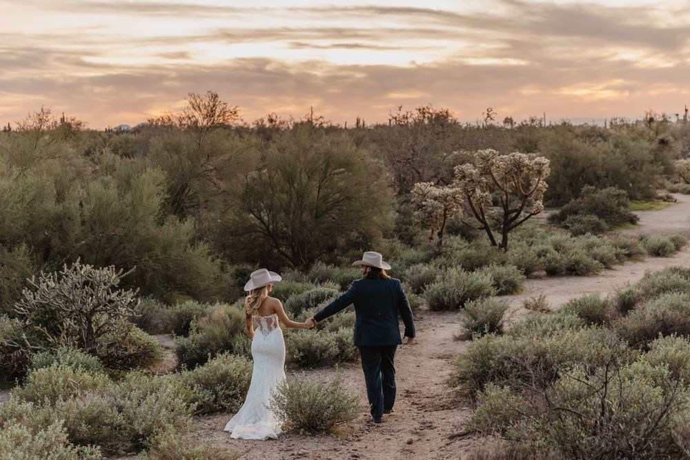 Bride and groom walk hand-in-hand along a desert path at sunset, surrounded by lush greenery and cacti.