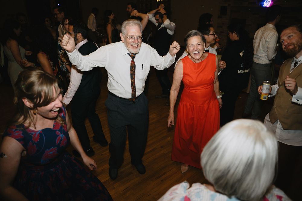 Elderly couple in festive attire dance enthusiastically with raised arms at wedding reception.