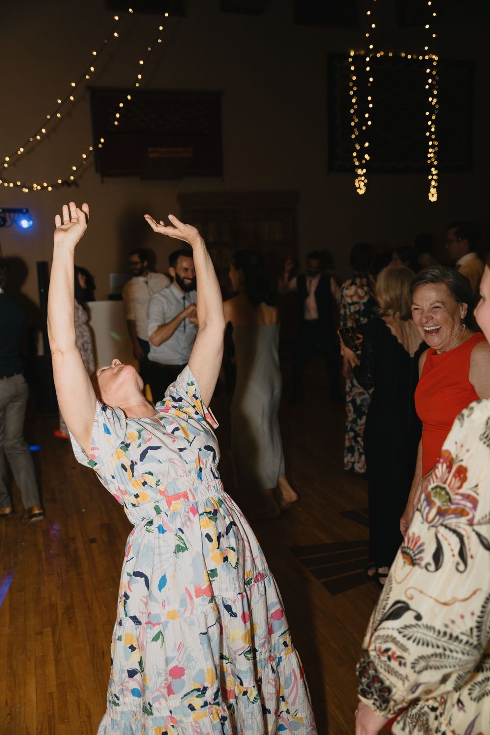 Wedding guest in floral dress joyfully raises arms while dancing at lively indoor reception.