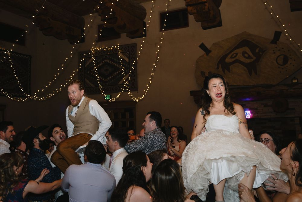 Bride and groom lifted by guests during lively wedding reception with twinkling fairy lights overhead.