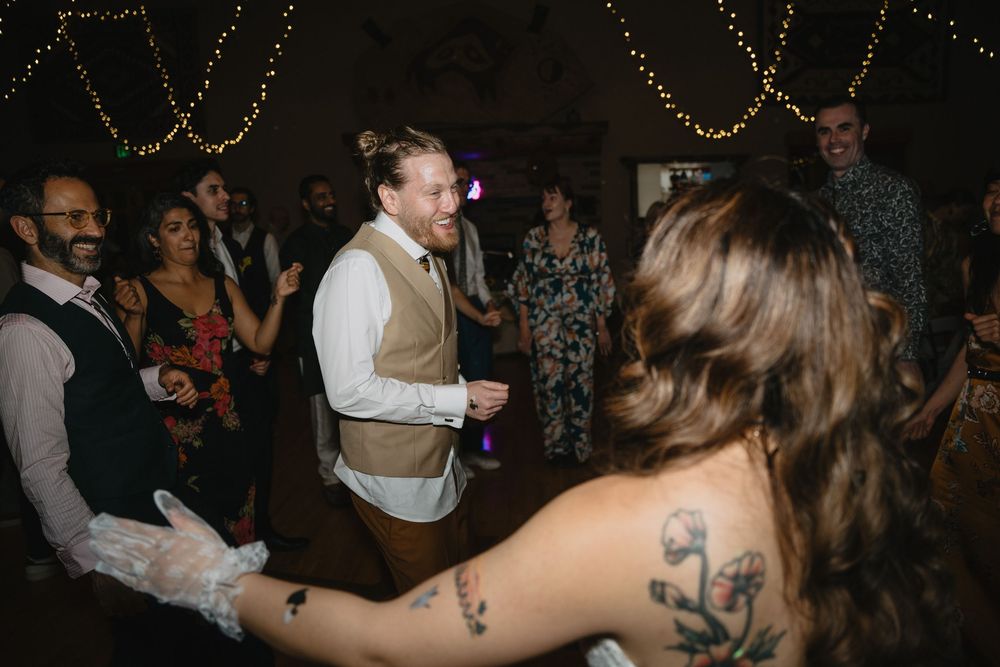 Groom in brown vest laughing while dancing with tattooed bride wearing lace gloves at reception.