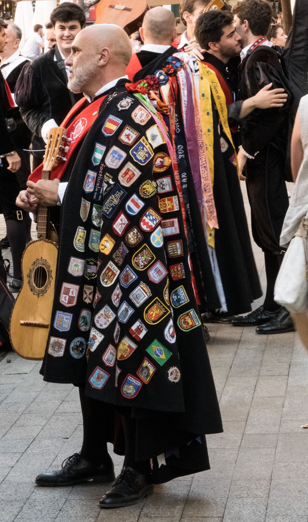 Man wearing a black cape covered in colorful heraldic patches and badges at a Spanish tuna street performance.