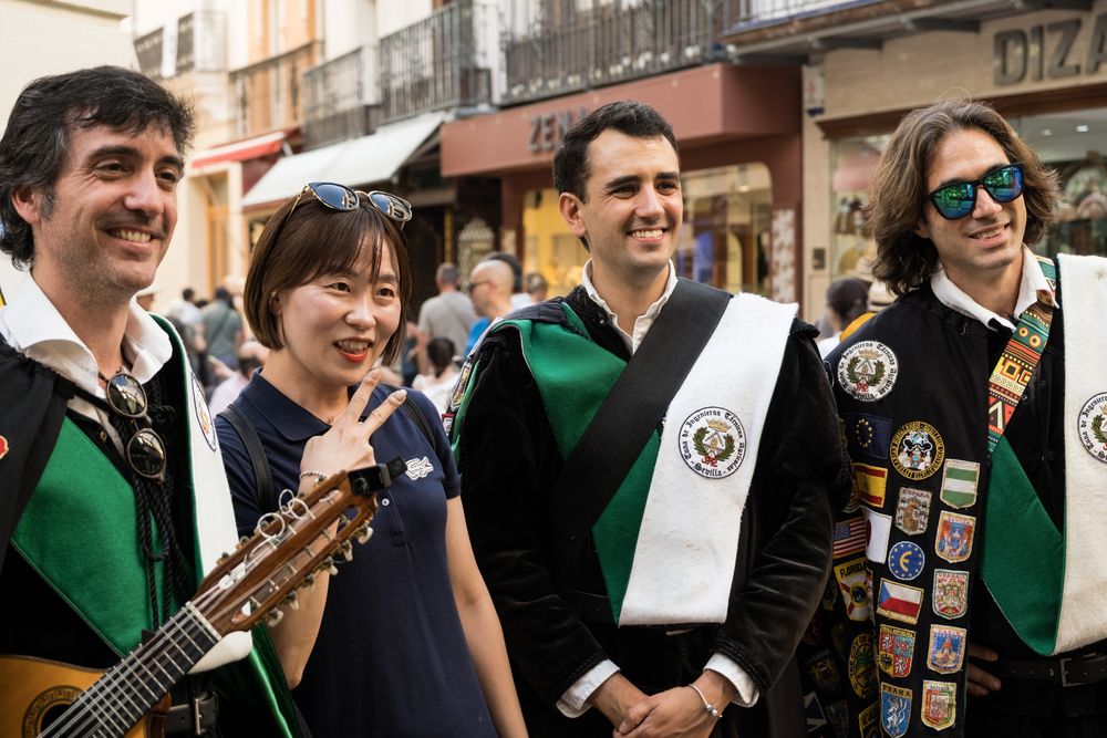 Four smiling people posing together with a guitar during a Spanish tuna musical street celebration in a historic city.