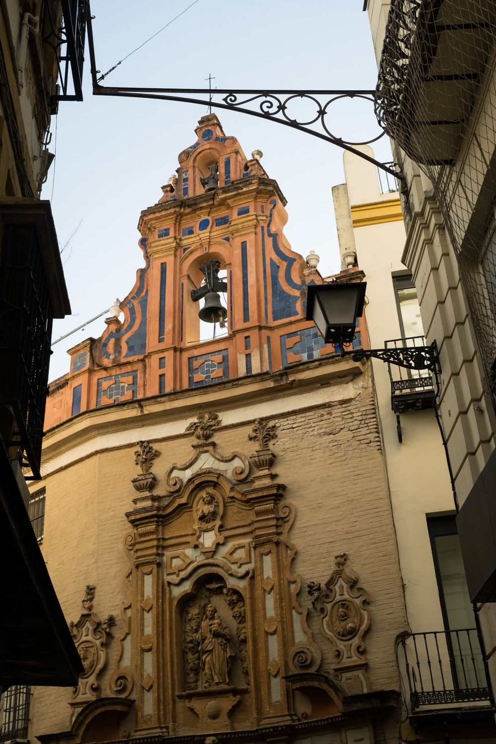 Baroque church facade with ornate bell tower, decorative stonework, and religious sculptures on a narrow Seville street.