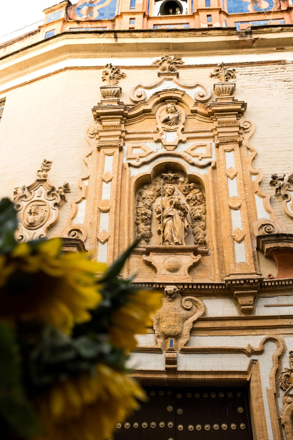 Ornate Baroque church exterior wall with detailed stone carvings, religious statue in niche, and sunflowers in foreground.