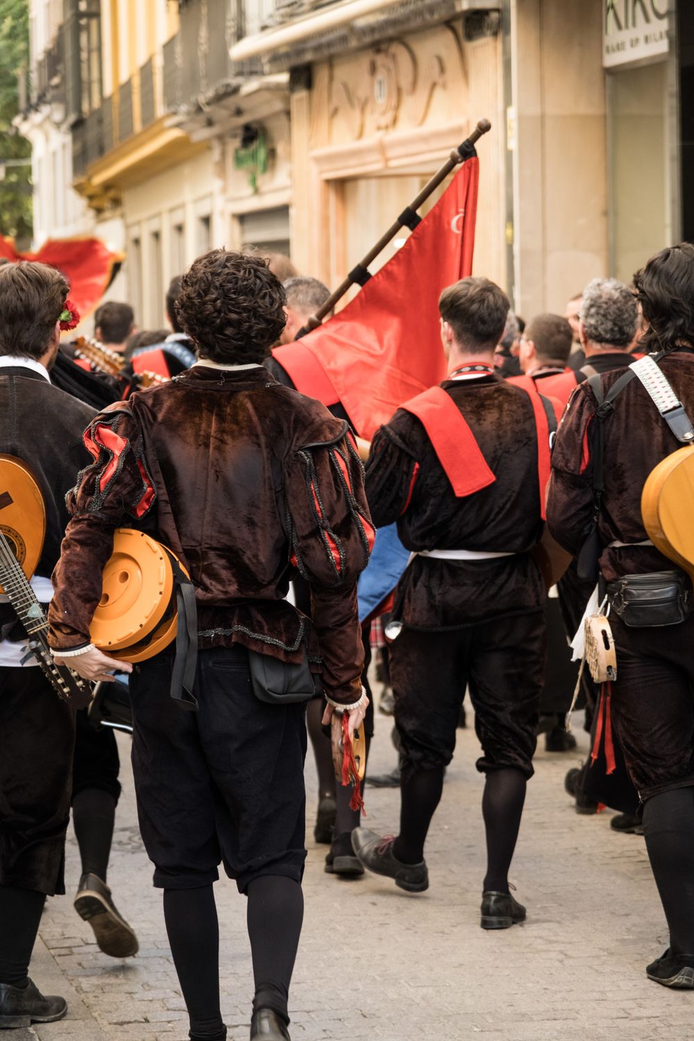 Rear view of Spanish tuna performers in black medieval costumes carrying red banners and instruments through city streets.