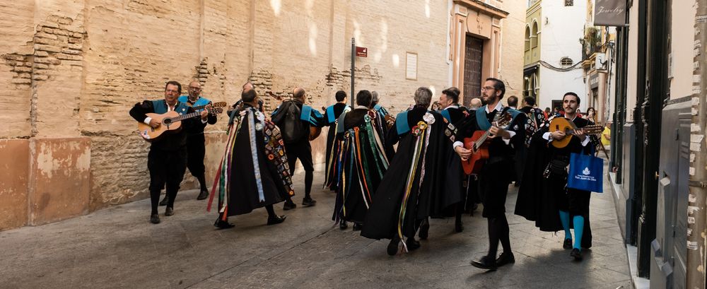 A Spanish tuna musical group in black capes with colorful ribbons performing in a narrow historic street.