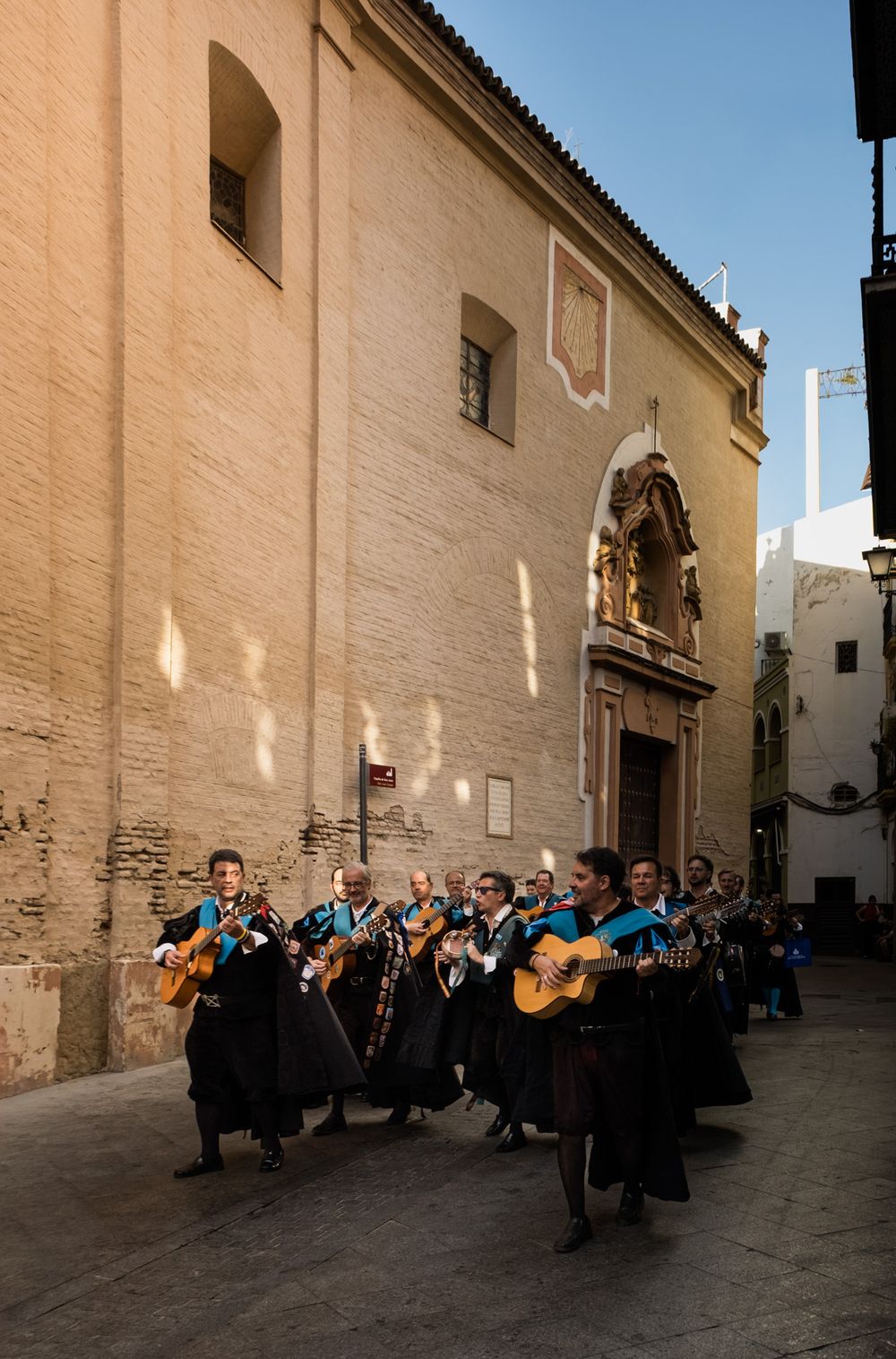 Traditional Spanish tuna musicians in black robes playing guitars outside a historic church building.