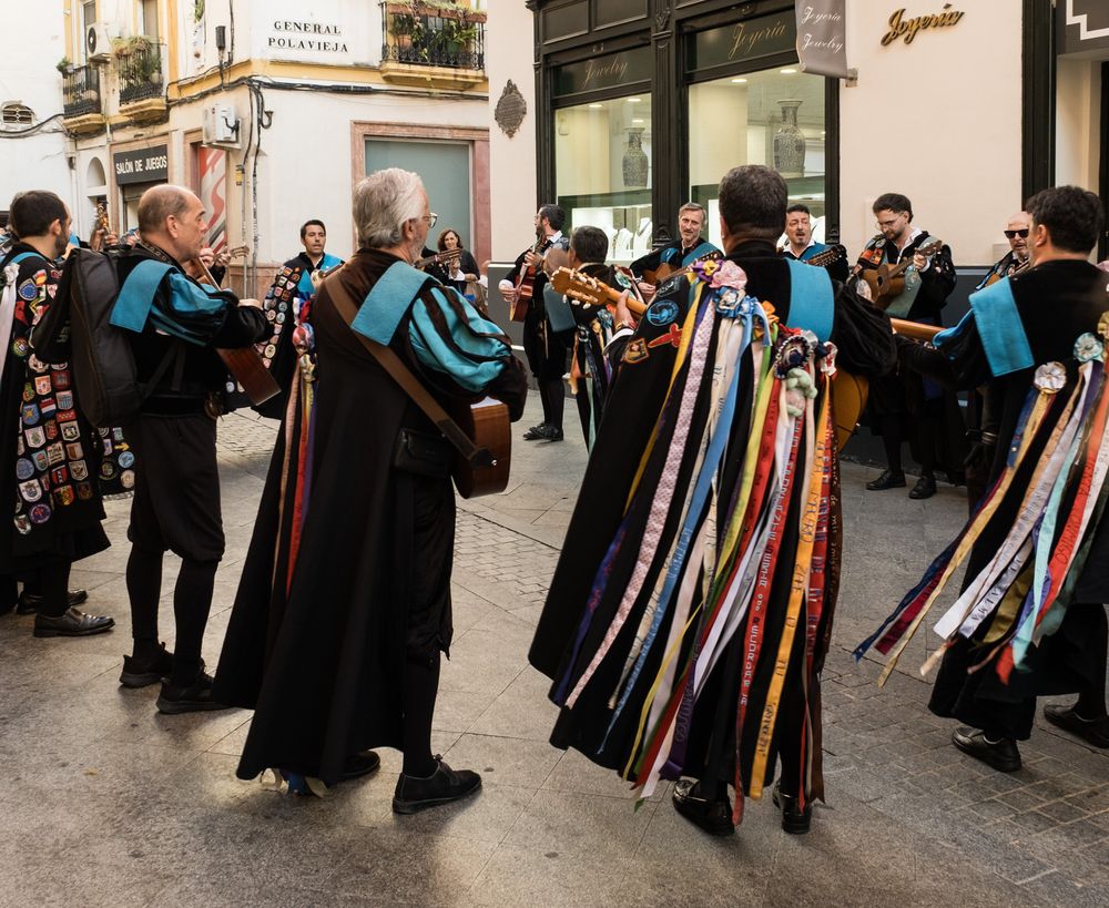 Spanish tuna students in black capes adorned with colorful ribbons playing instruments in a town square.