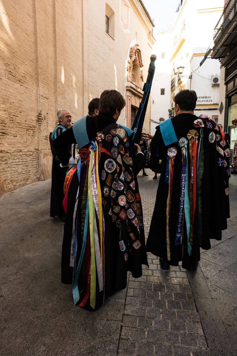 Rear view of Spanish tuna members in black capes decorated with colorful patches and ribbons walking through narrow streets.