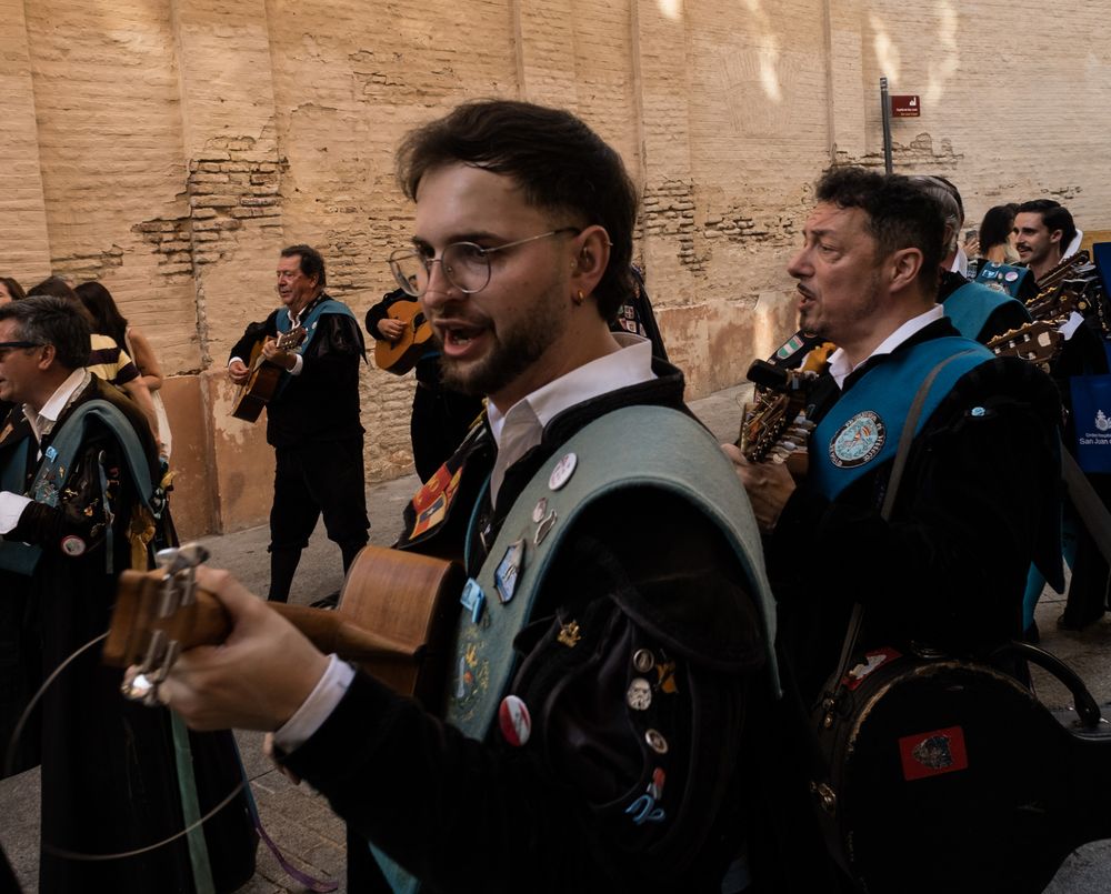 Young Spanish tuna musician in decorated black cape playing a stringed instrument in a historic street.