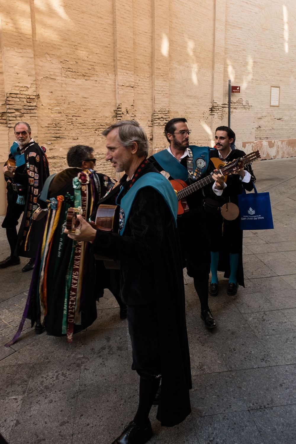 Spanish tuna musicians in black academic robes with teal sashes playing guitars in a sunlit alley.