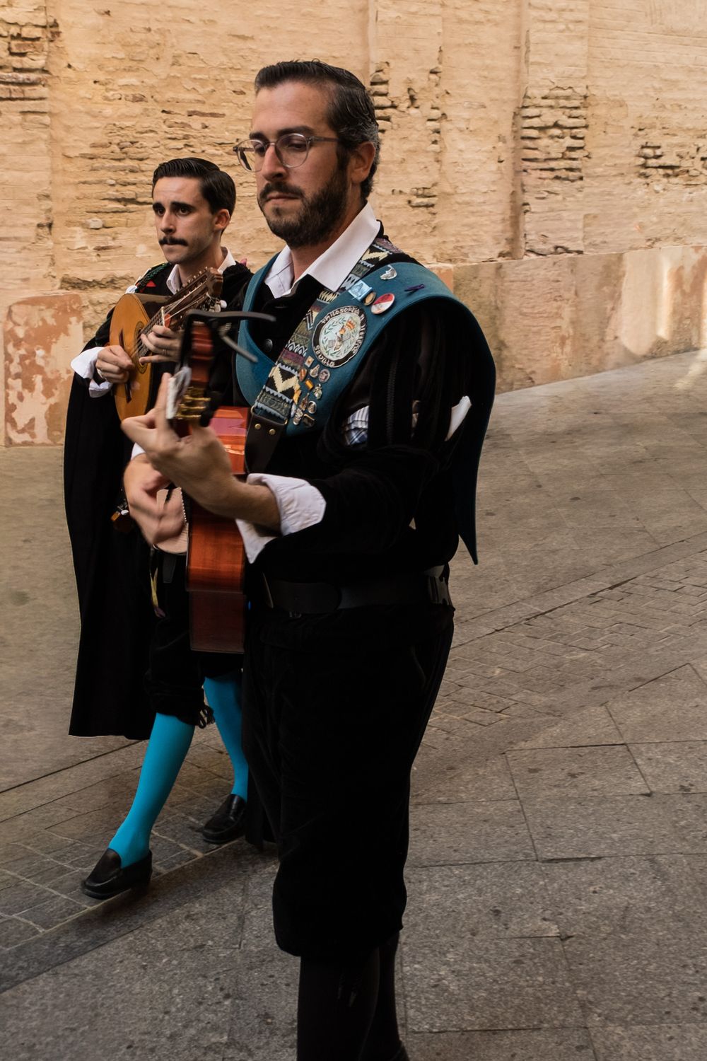 Two Spanish tuna musicians in black robes, one wearing teal tights, playing guitar outdoors.
