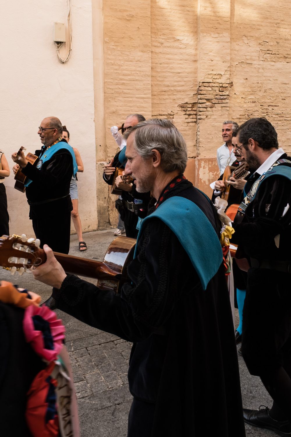 Spanish tuna musicians in black robes with teal sashes playing guitars against a weathered stone wall.