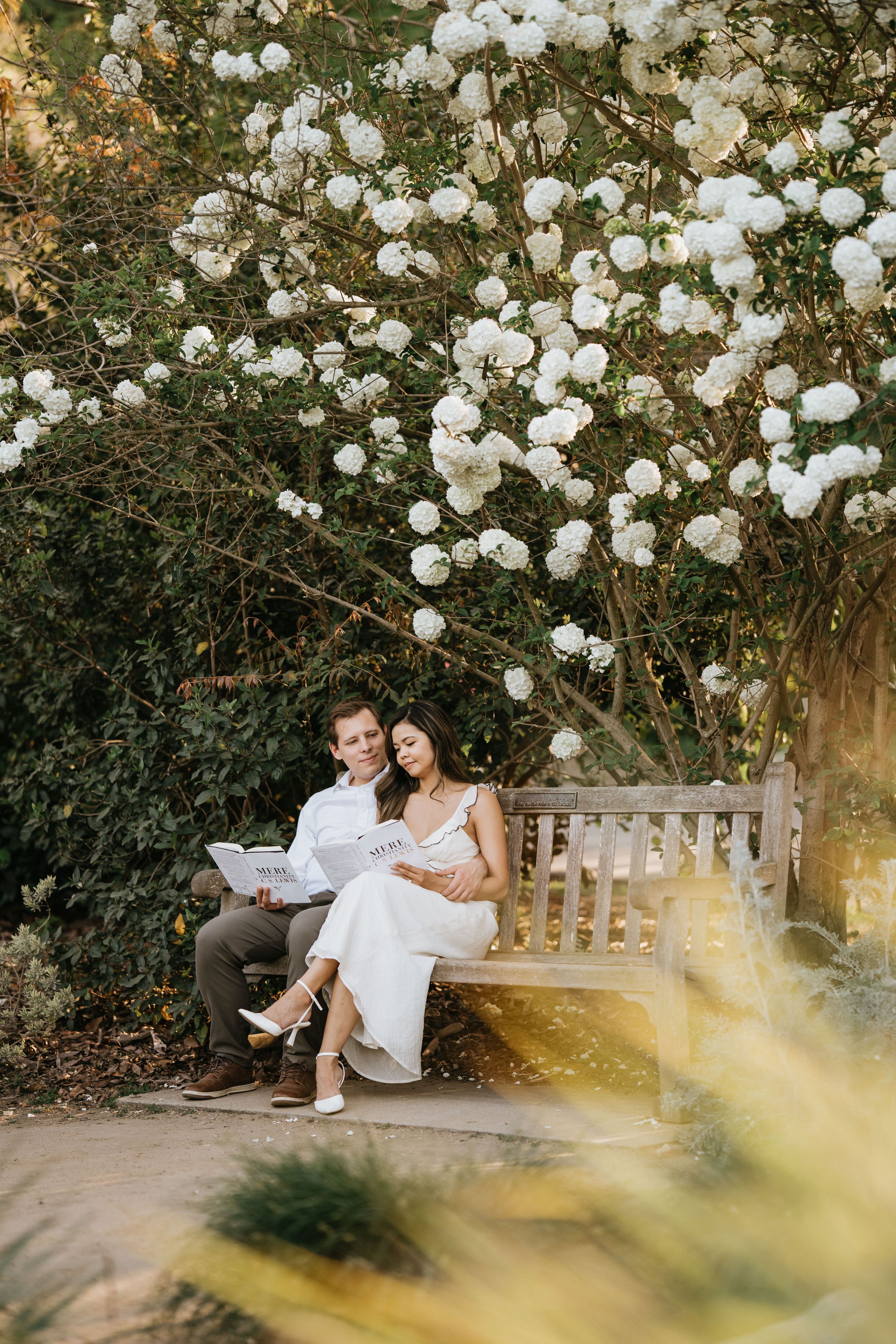 Mariel and Jeff reading together on a garden bench beneath blooming white flowers.