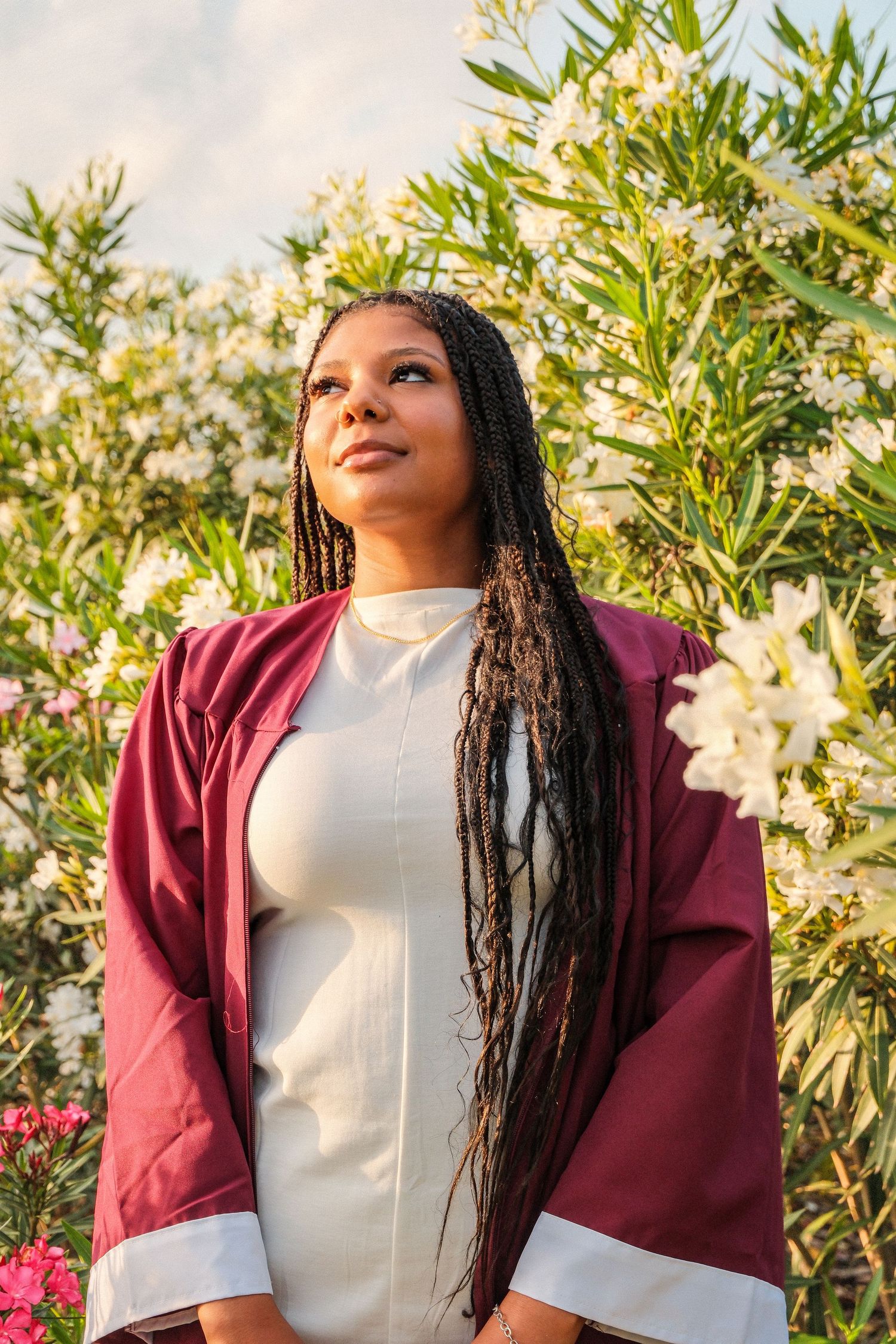Graduate woman in maroon cap and gown standing before blooming white flowering bushes.
