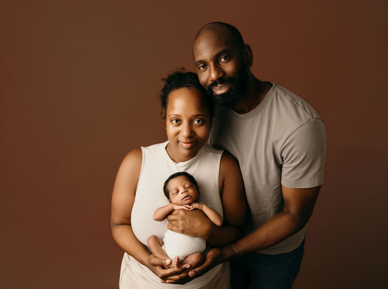 A joyful family portrait of parents holding their newborn baby against a warm brown studio background.