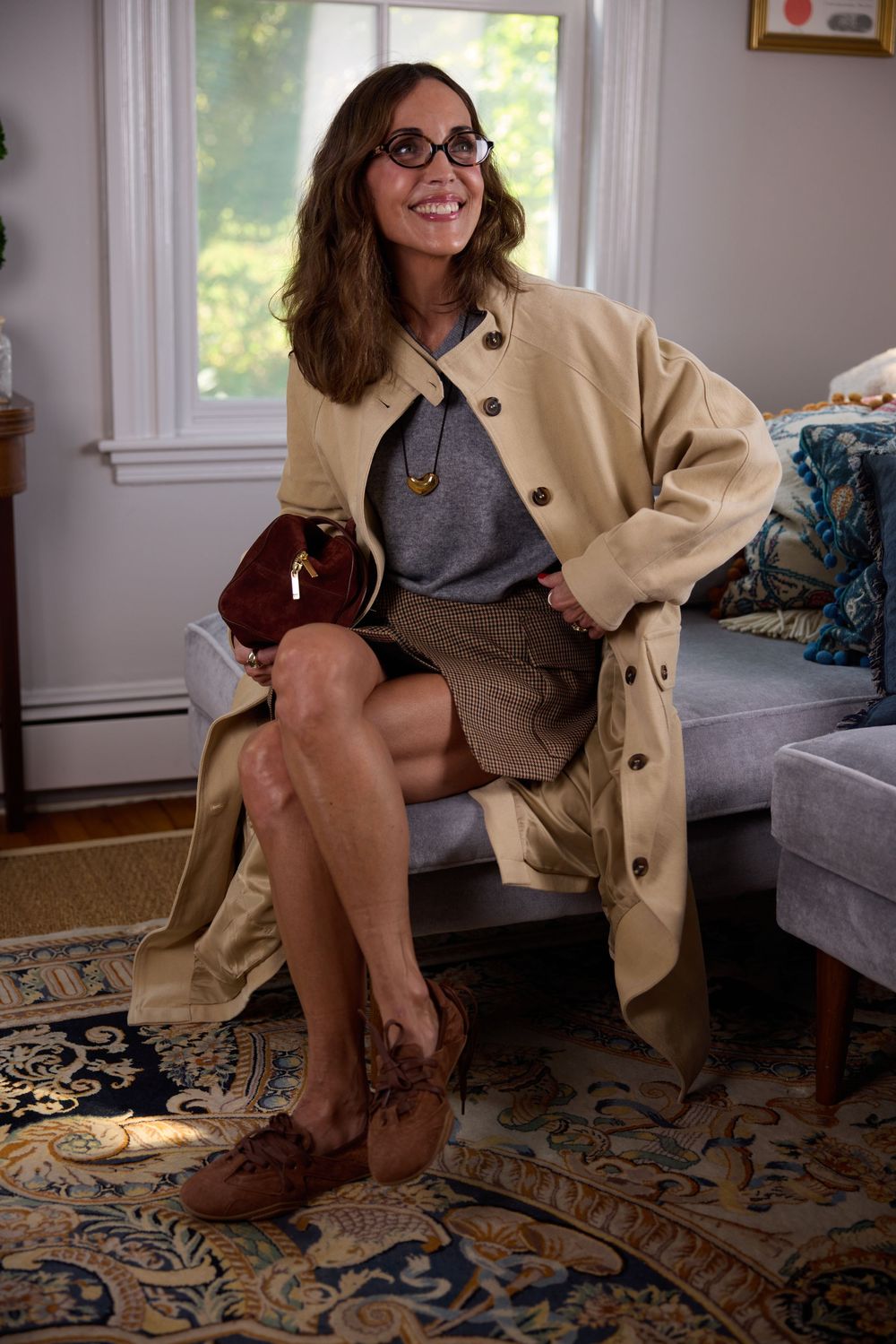 Woman in beige trench coat and plaid skirt holding brown bag while seated in living room.