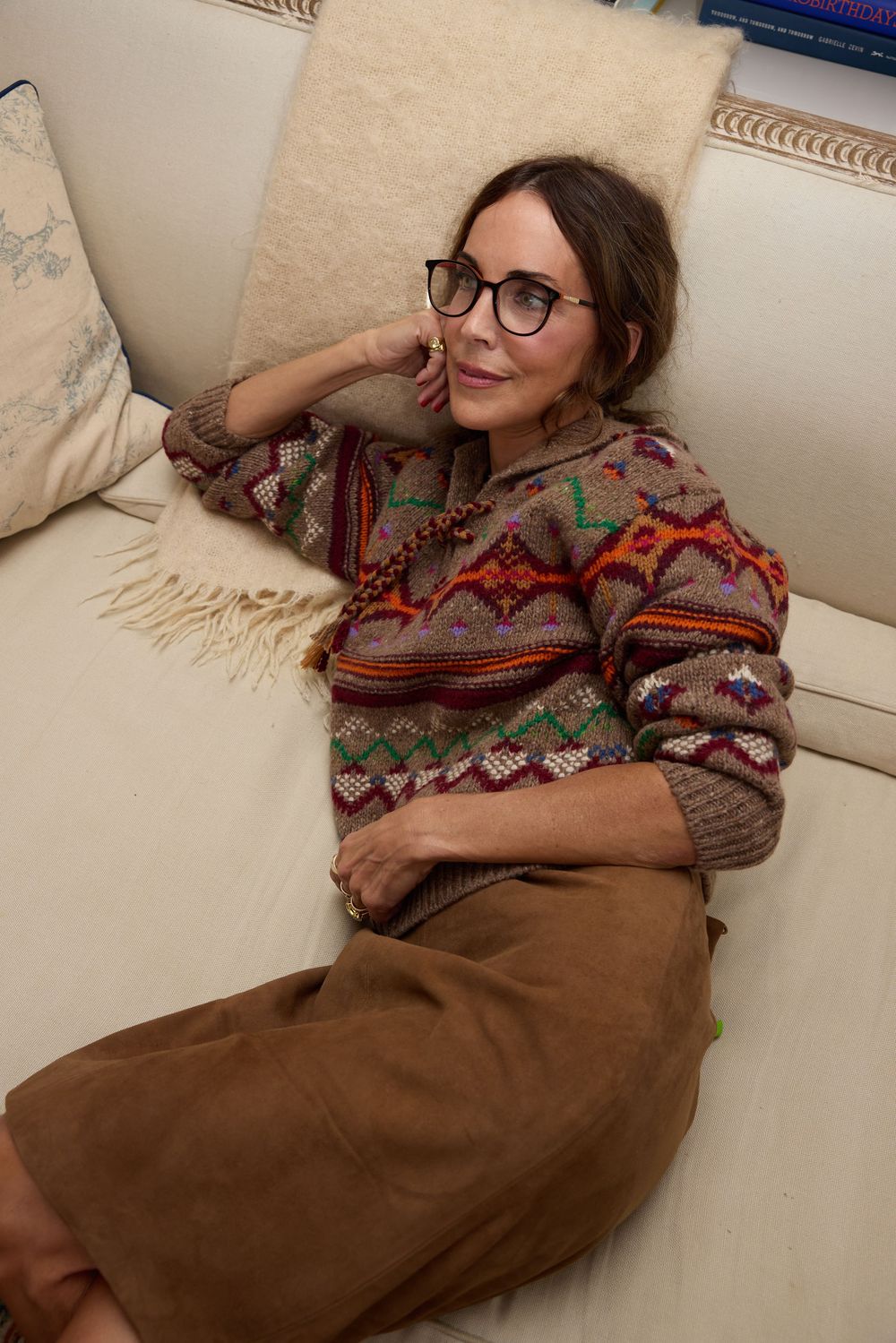 Smiling woman in multicolor patterned sweater and brown skirt relaxes on white sofa cushions.