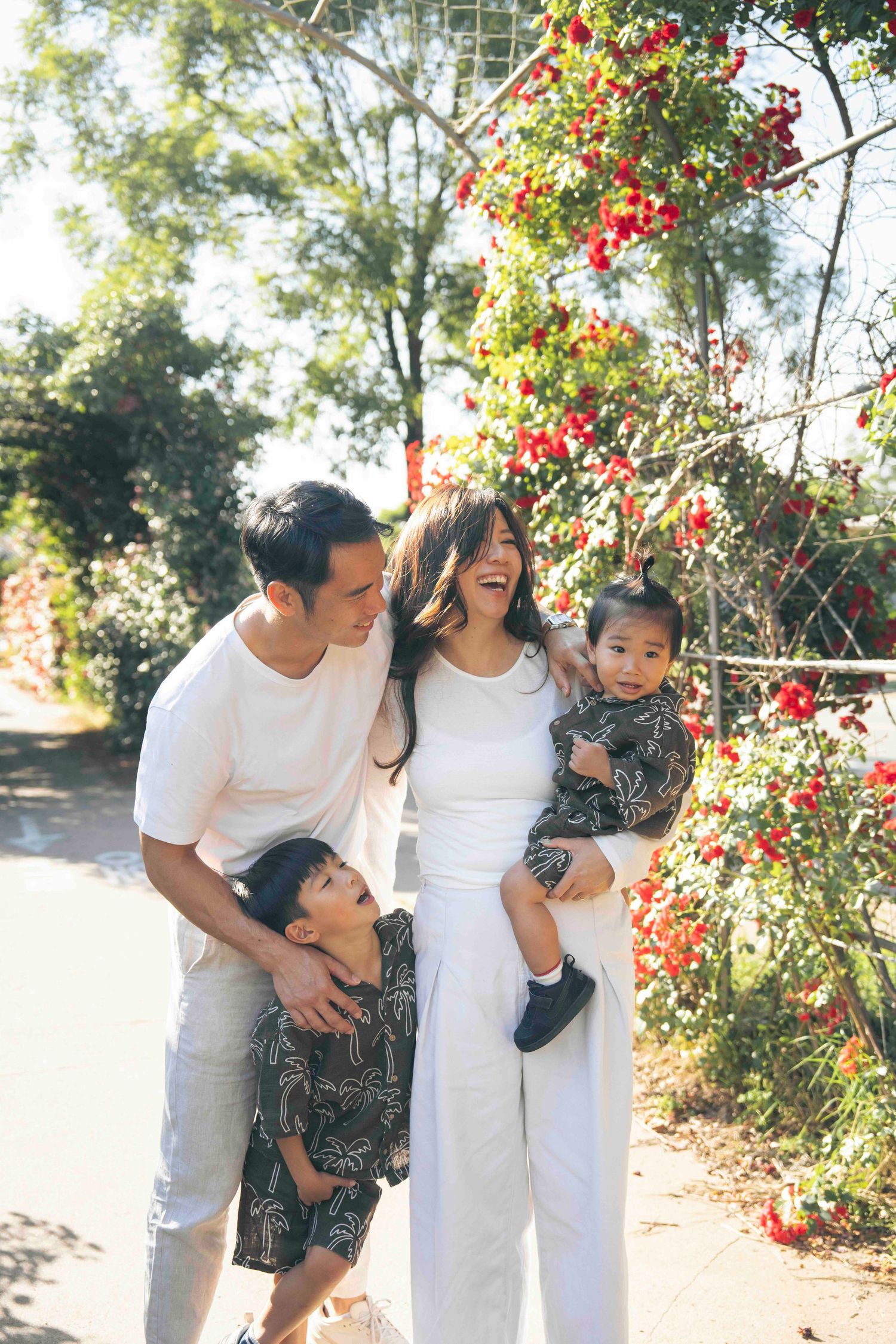 Happy Asian family of four posing outdoors near red flowering vines on a sunny day.