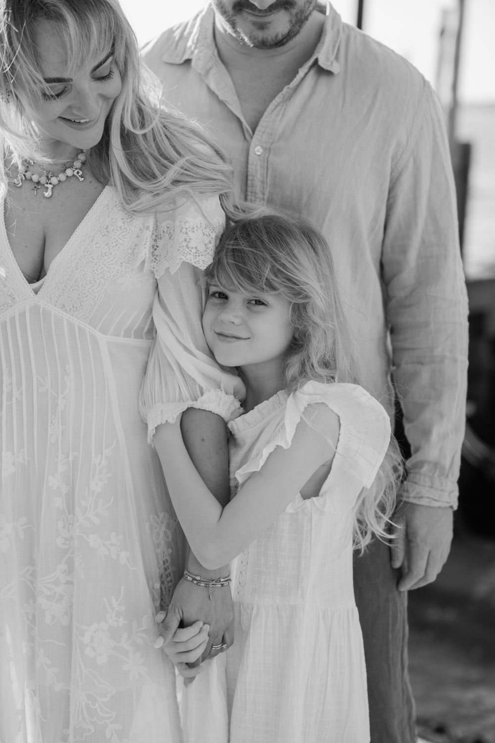 Black and white family portrait of parents and young daughter in white dresses sharing a tender embrace outdoors.