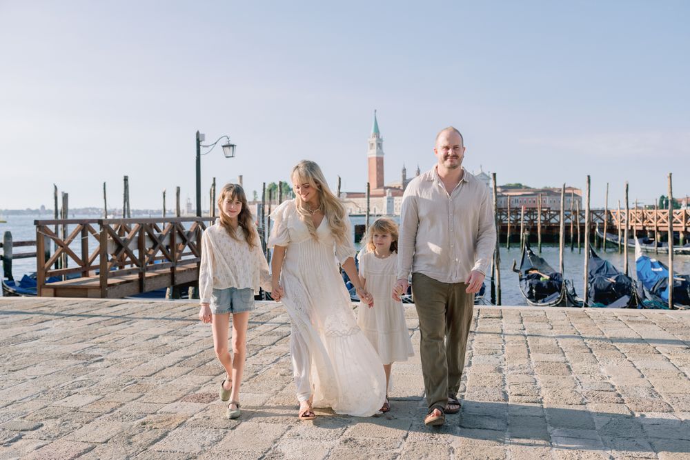 Family of four walking along Venice waterfront with gondolas and San Giorgio Maggiore church in background.
