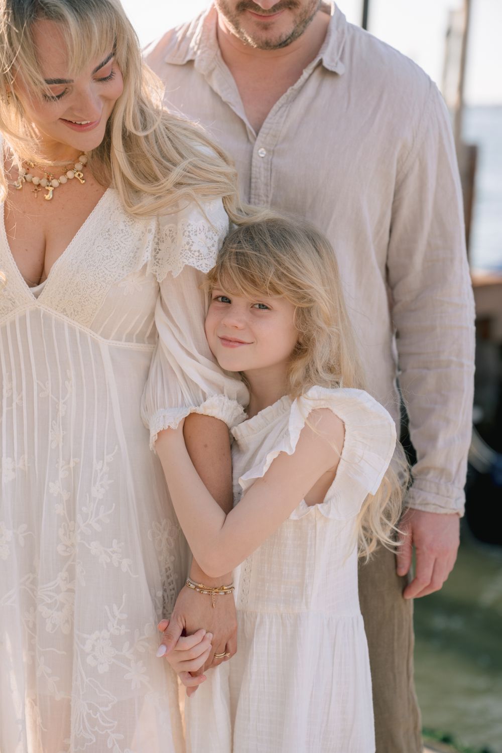 A blonde family in white and beige linen outfits, with a young girl hugging her mother outdoors.