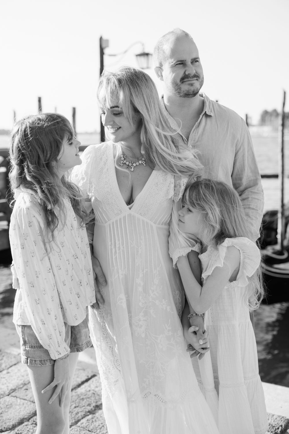 Black and white family portrait of parents and two young daughters posing together outdoors near a waterfront.