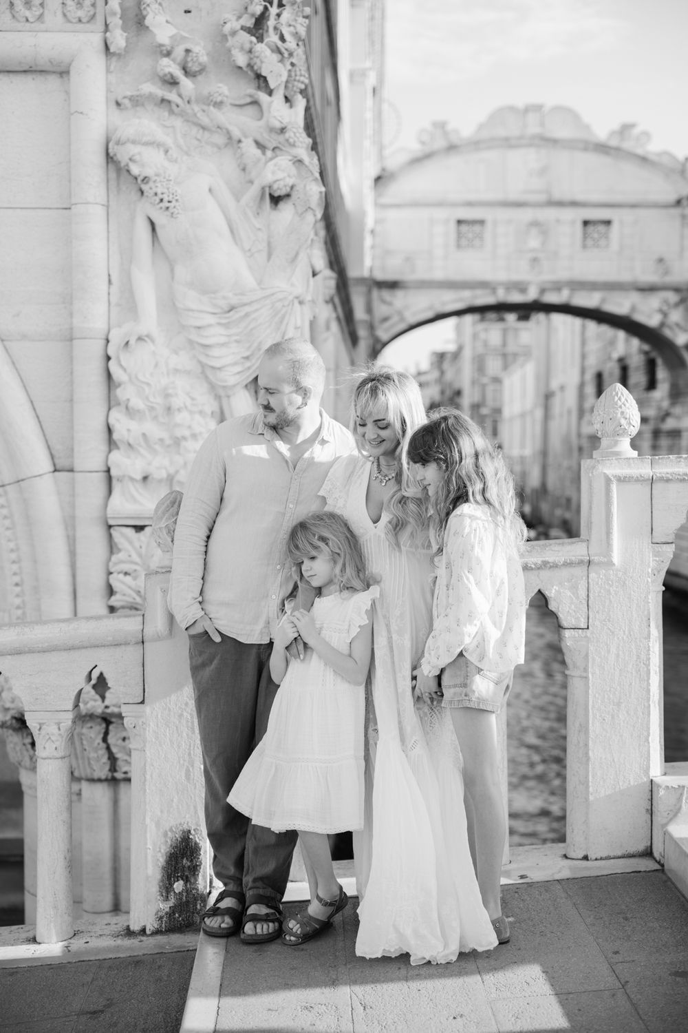 Black and white family portrait in Venice, parents and two daughters posing by ornate stone bridge architecture.