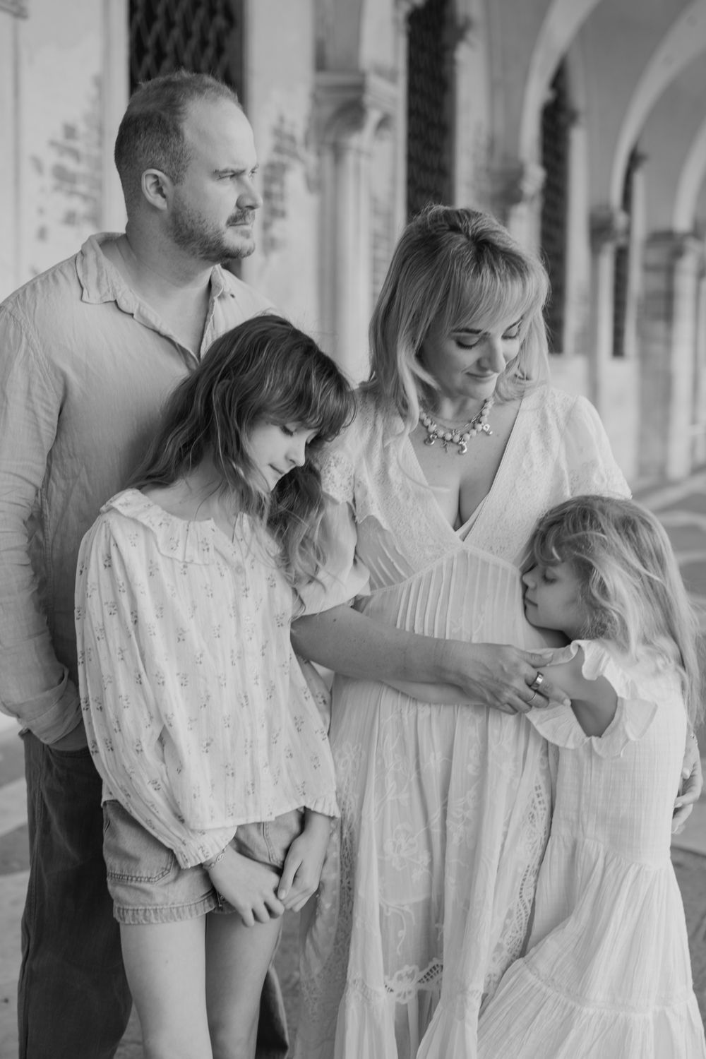 Black and white family portrait of parents with two young daughters in white dresses posing in an elegant archway.