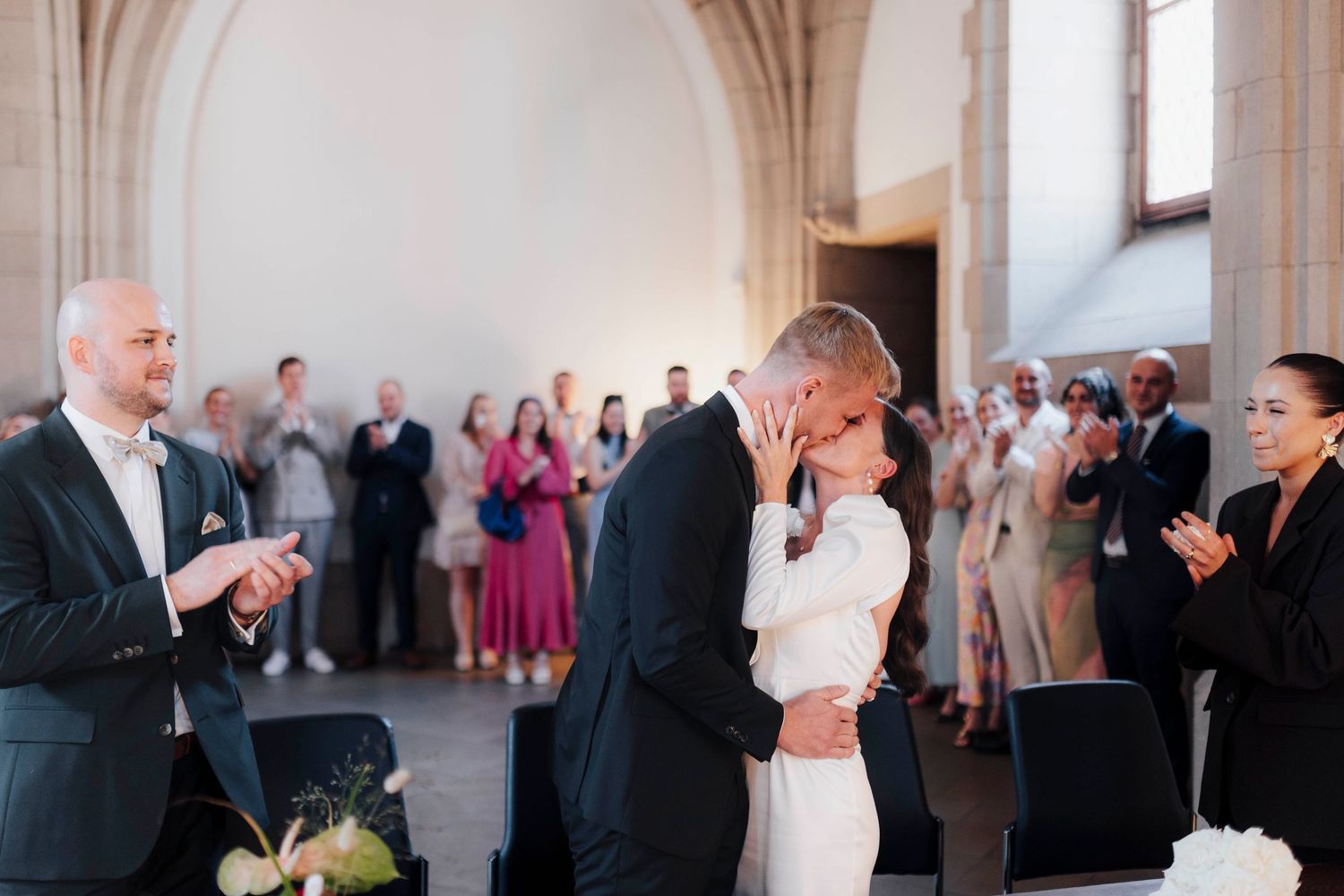Bride and groom share first kiss at elegant wedding ceremony while guests applaud in bright architectural venue.