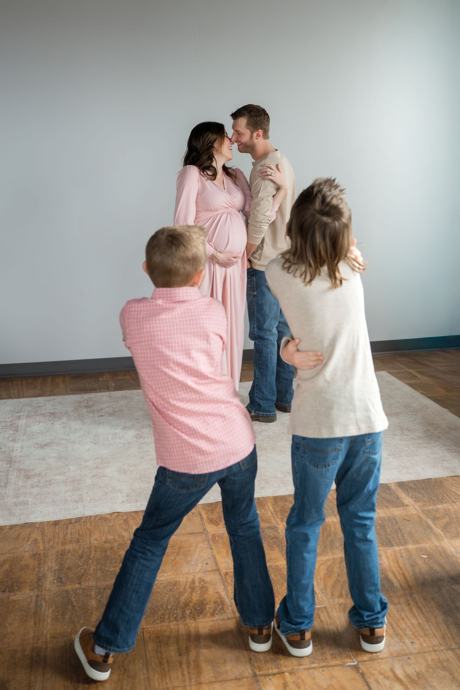 Pregnant woman and man kissing while two children hug them during a family maternity photo session indoors.
