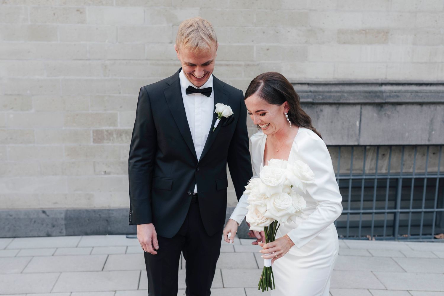 Bride in white gown holding bouquet and groom in black tuxedo share a joyful moment outdoors on their wedding day.