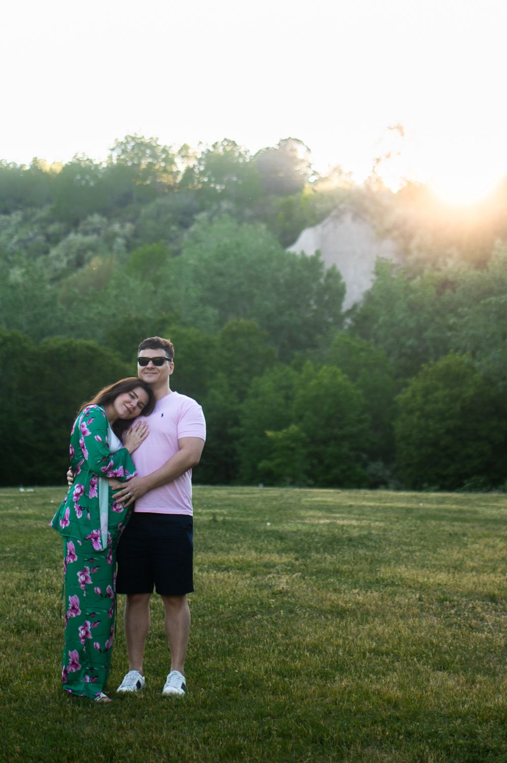 Couple embracing at golden hour in a green park, woman in floral dress leaning on mans shoulder.