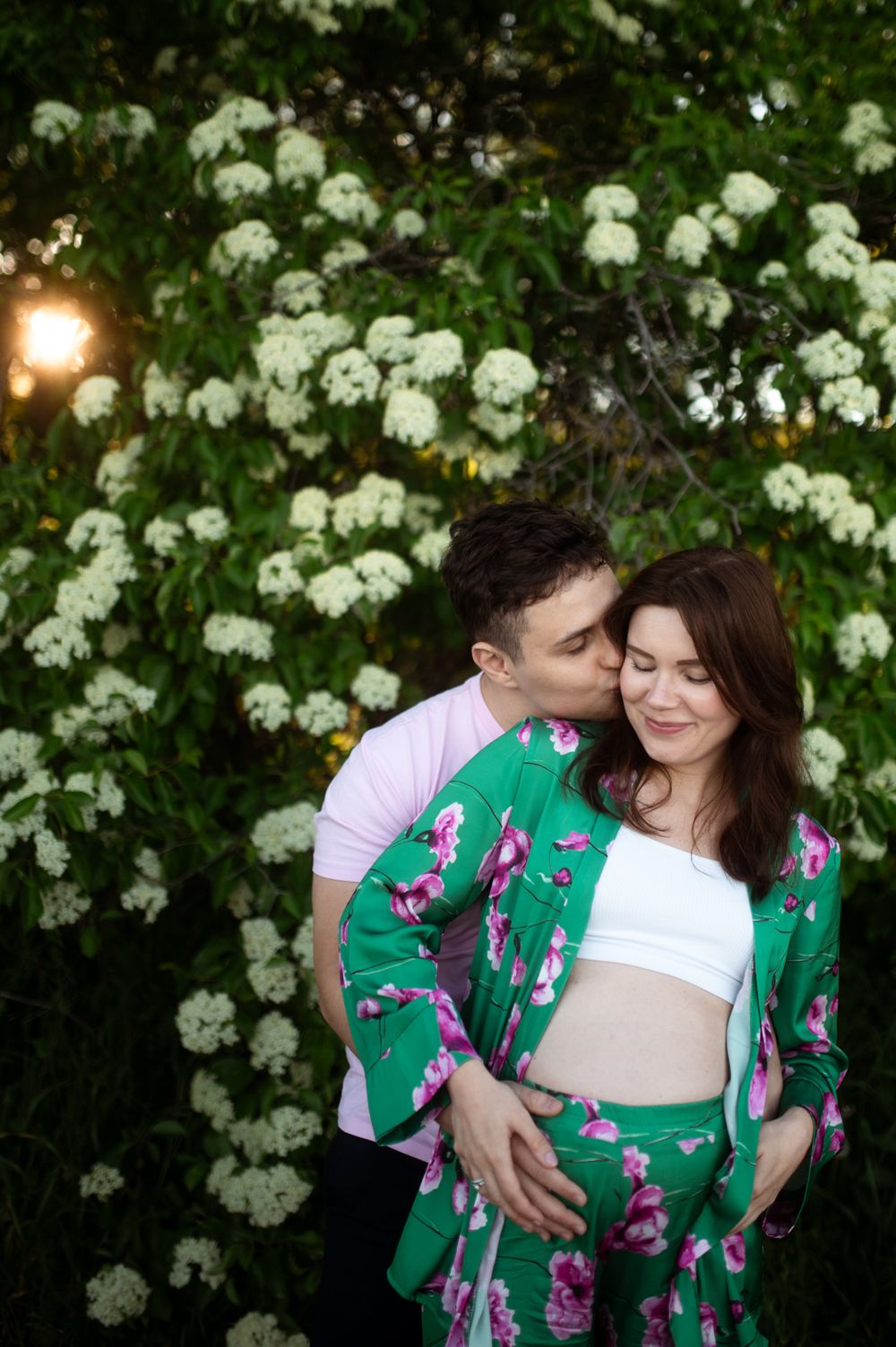 Man kissing pregnant woman near white flowering bushes, she wears a green floral outfit.