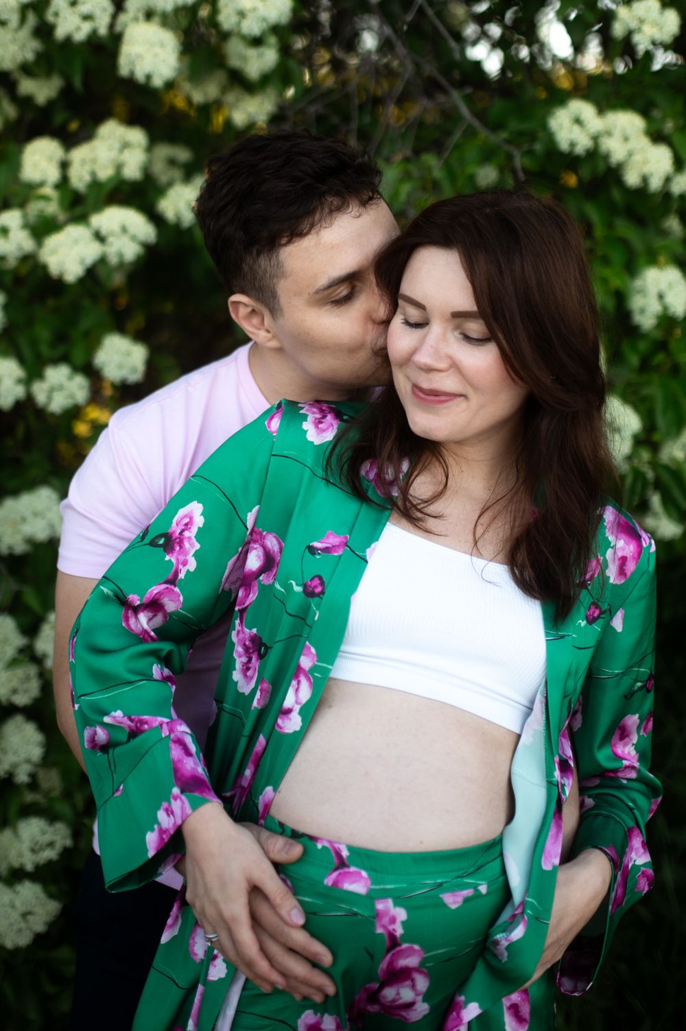 Couple sharing a tender moment outdoors, pregnant woman in green floral suit with white crop top, man in pink shirt kissing her cheek.