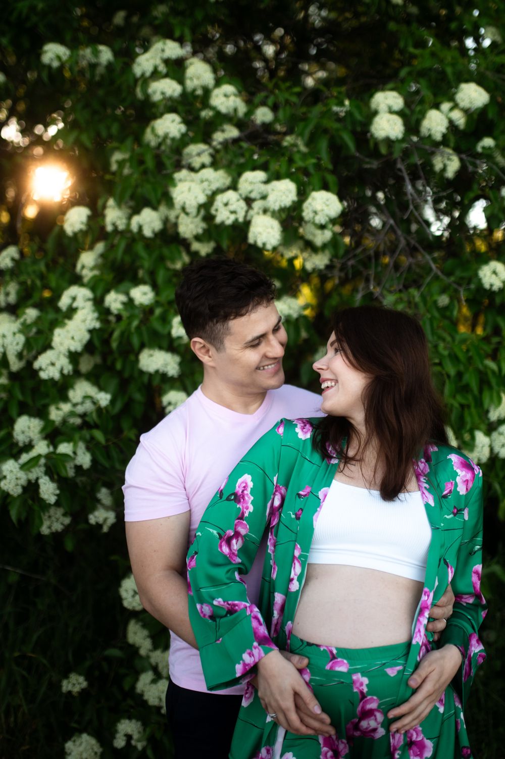 Smiling couple embracing in front of white blooming flowers, woman in green floral maternity outfit.