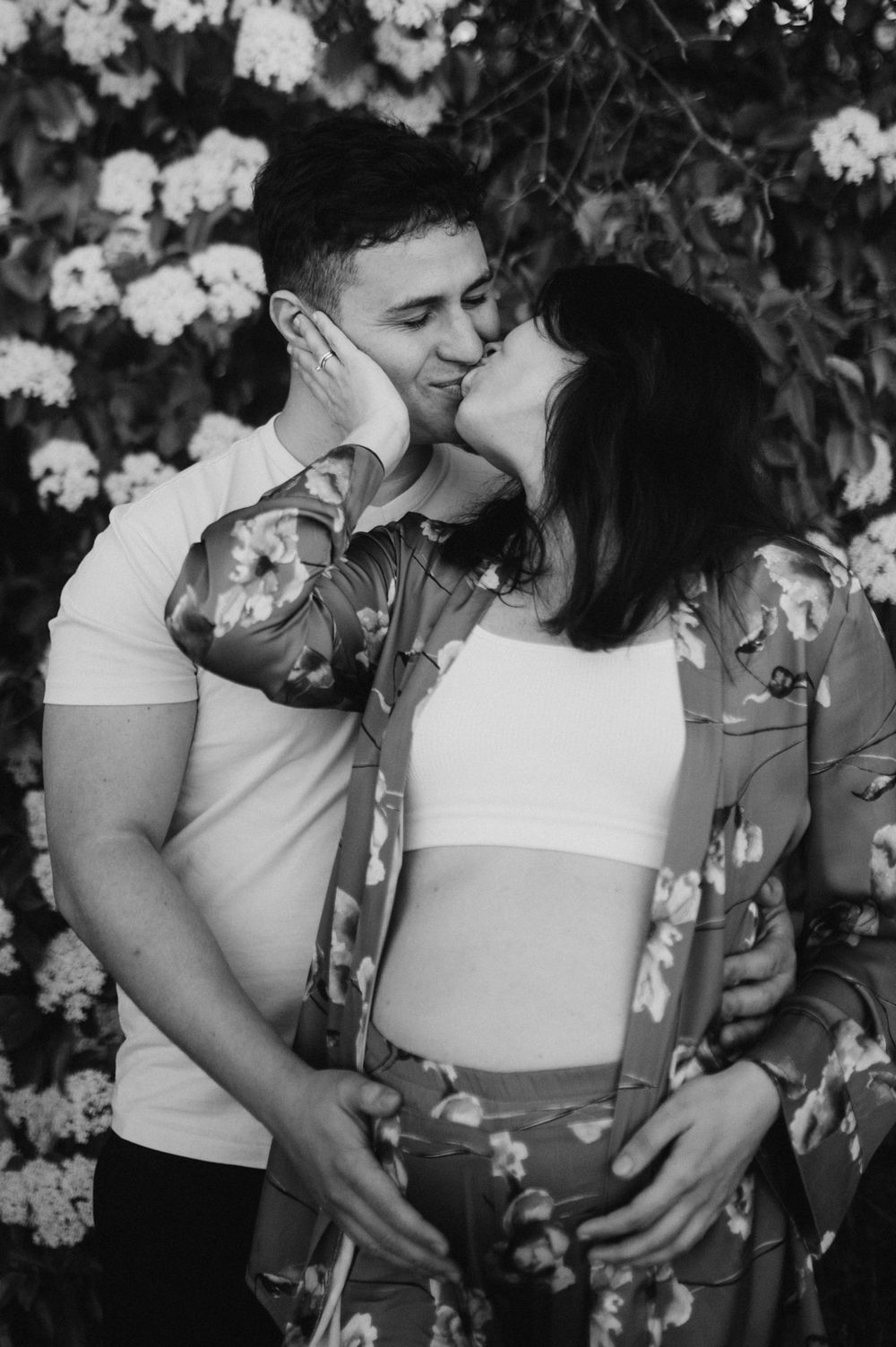 Black and white photo of couple kissing near white flowering bushes, maternity photoshoot.