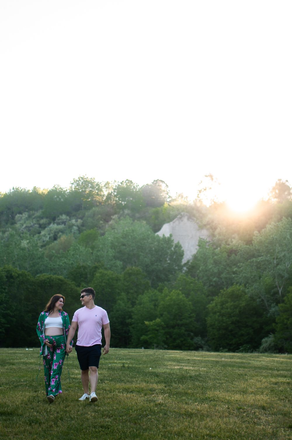 Couple holding hands walking through a sunlit green field with trees and golden sunset behind them.