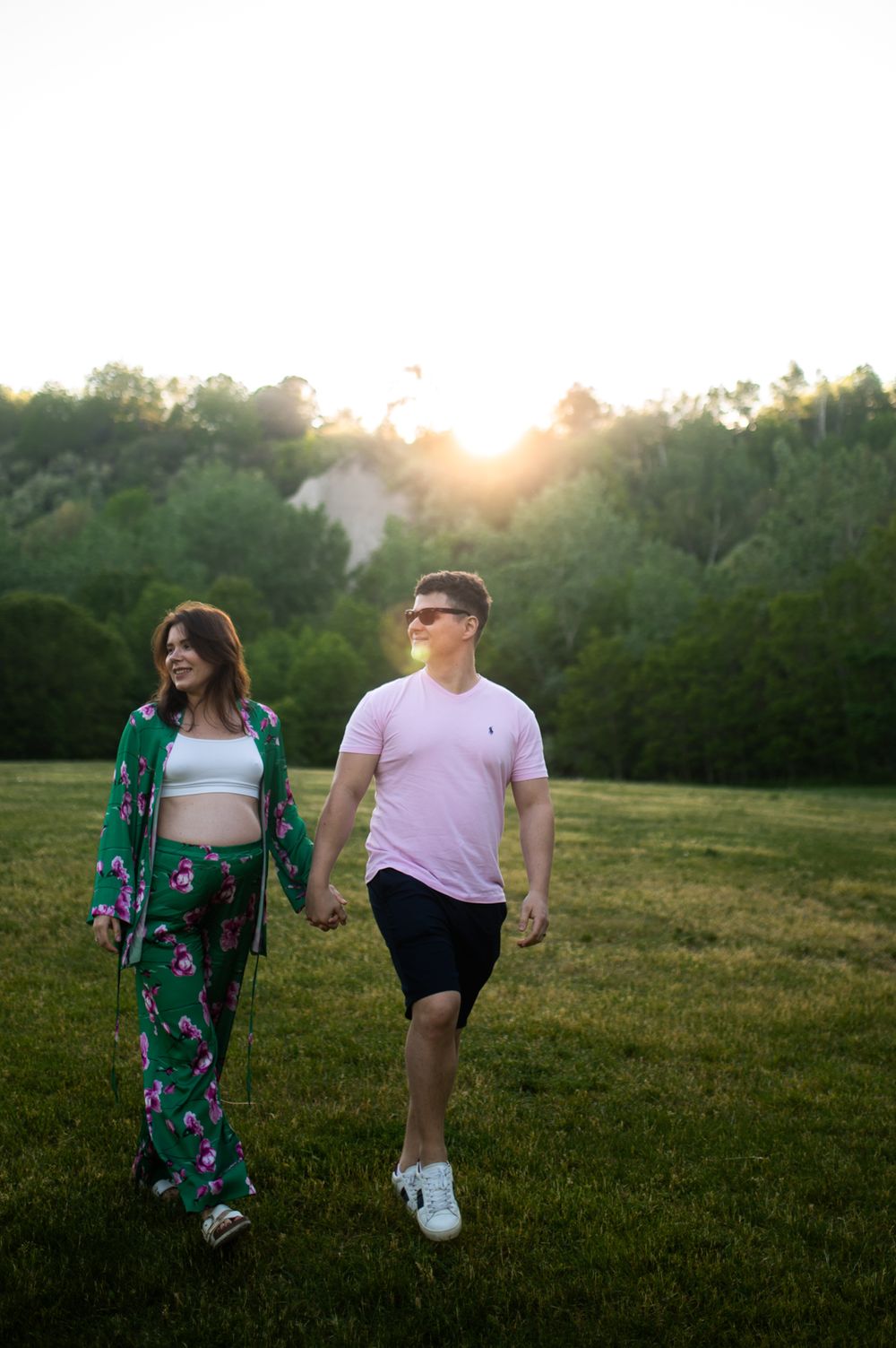 Couple holding hands walking in a sunlit green field at golden hour, woman in floral outfit, man in pink shirt.
